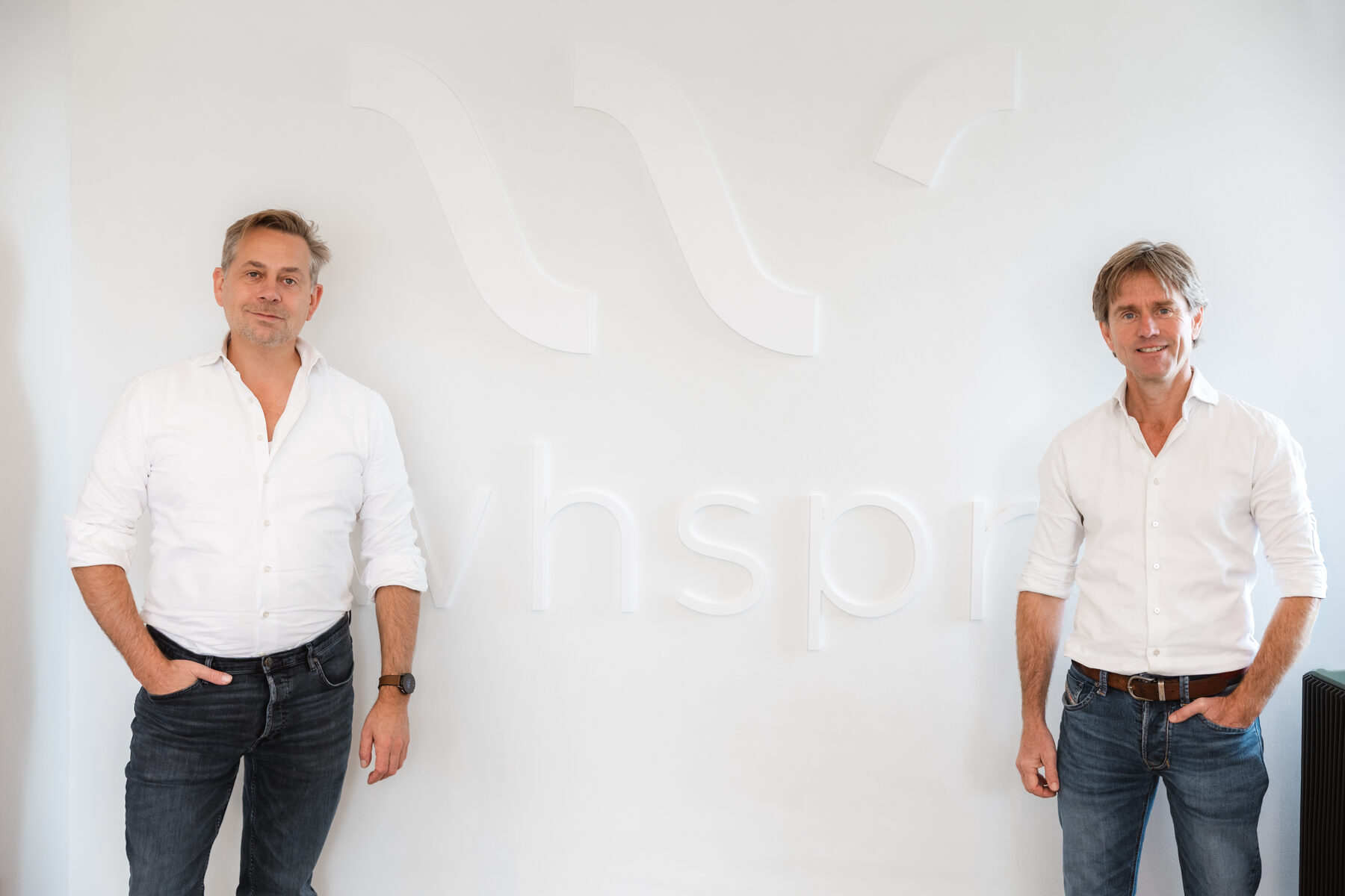 Twee oprichters in witte shirts voor een witte wand met het grote Whspr-logo